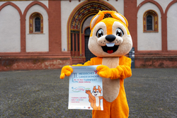 Das Maskottchen Freddy hält ein Plakat vom Landeskinderturnfest in den Händen.