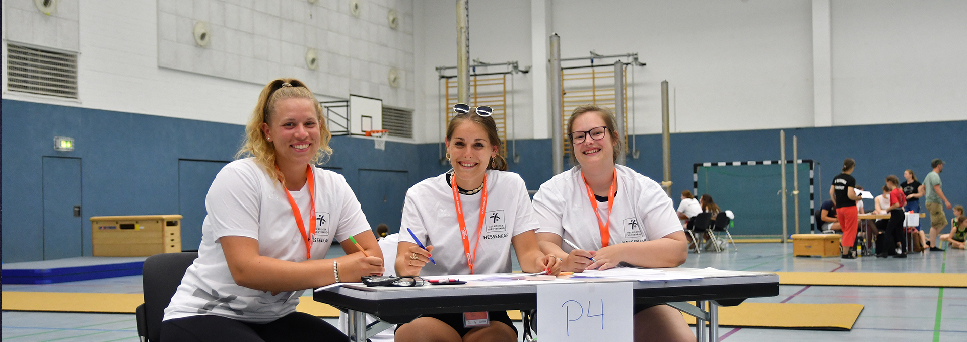 Drei junge Frauen sitzen an einem Kampfrichtertisch in einer Turnhalle und lächeln.